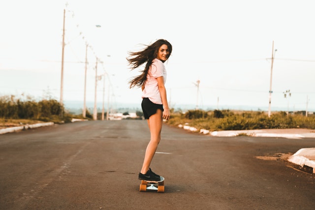 žena s dlouhými hnědými vlasy venku jede po ulici na skateboardu